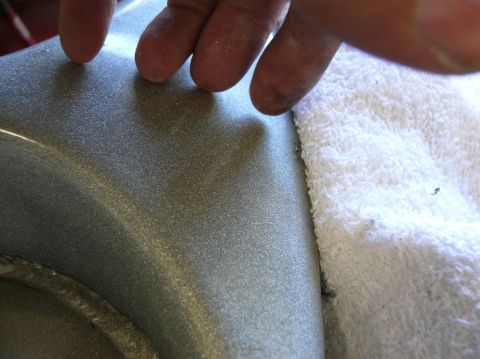 Close-up of metallic gray car part with glittery finish, fingers touching surface, white towel nearby