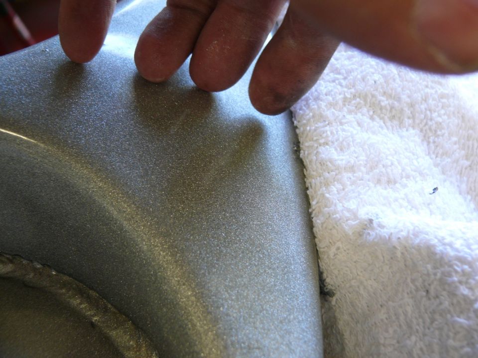 Close-up of metallic gray car part with glittery finish, fingers touching surface, white towel nearby