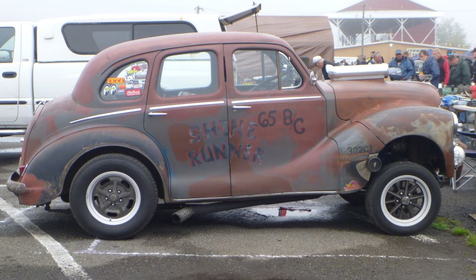 Rat rod car with custom paint, large intake, and aftermarket wheels. Text on door reads 'SHINE 65 BG RUNNER'