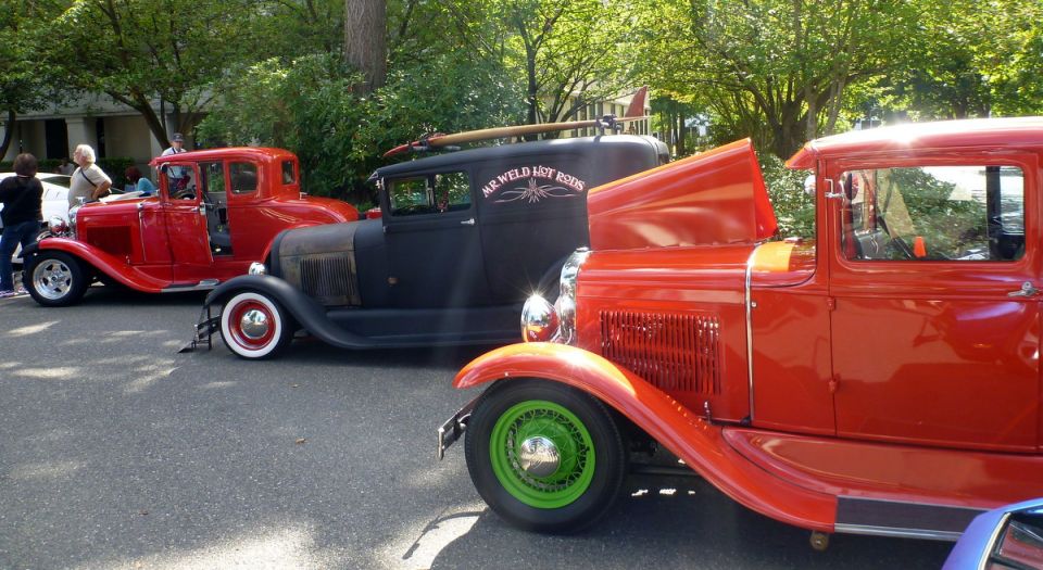 Three hot rod cars, two red and one black, parked on a street. Black car has 'MR. WELD HOT RODS' text and a surfboard on roof