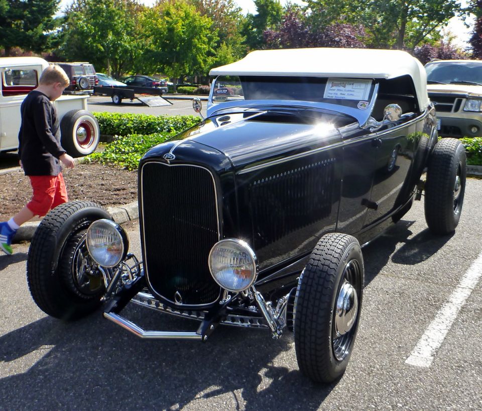 Black hot rod convertible with a white top, parked next to a boy. Another car is visible in the background