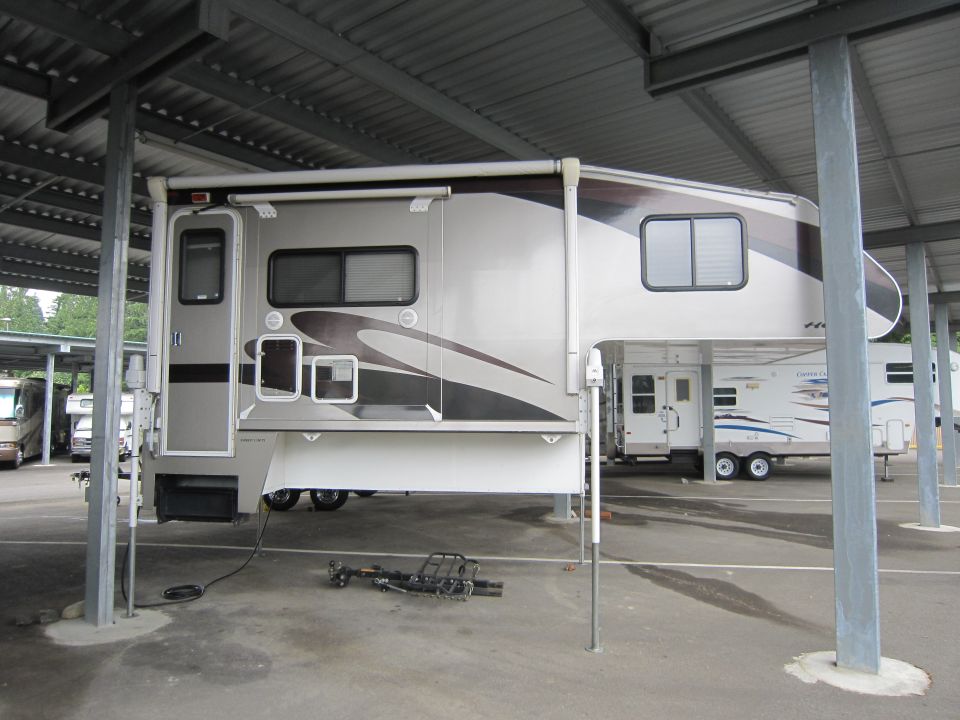 Tan slide-in truck camper on jack stands under a metal carport with a hitch bar on the ground