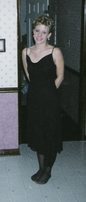 1977 Ford Mustang II - Woman in a black dress and pantyhose stands indoors with hands behind her back.