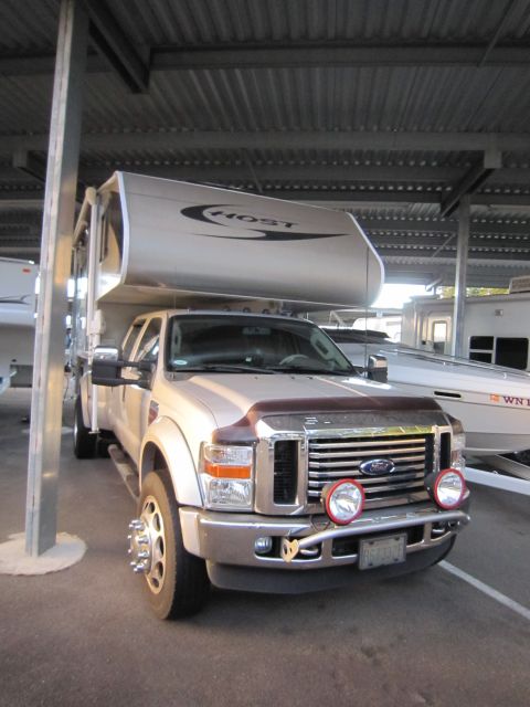 2008 Ford F-450 - A Ford truck with a camper shell is parked under a covered structure next to a boat.