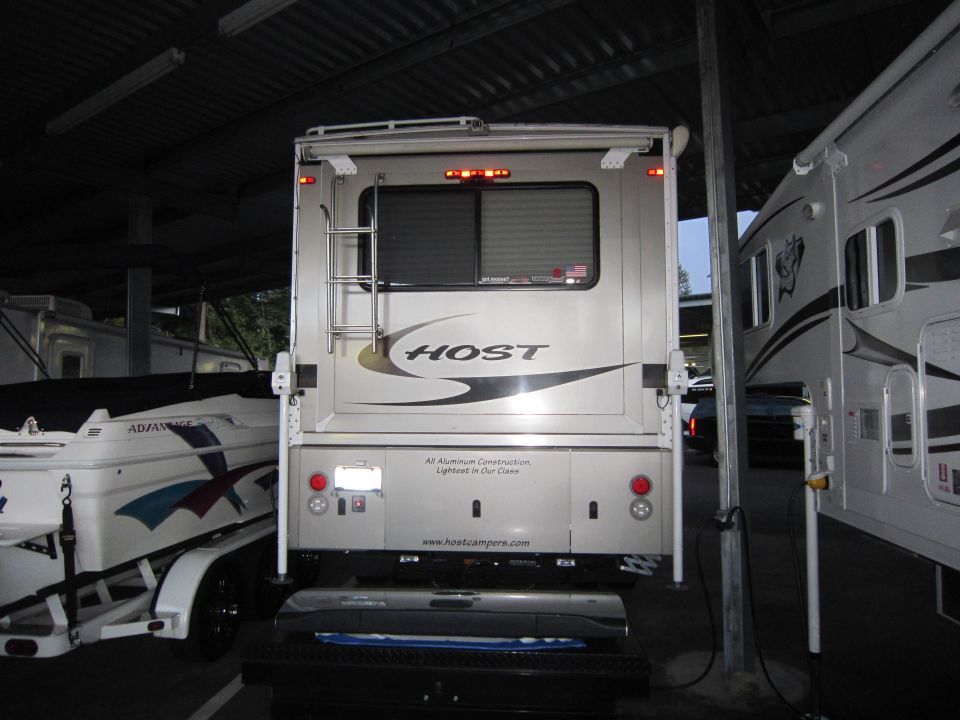 Rear view of a Host camper truck bed with a ladder and window. Another camper is to the right, and a boat on a trailer is to the left