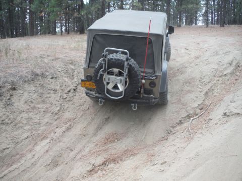 2005 Jeep Wrangler - A tan Jeep Wrangler with a soft top navigates an off-road trail, showing its rear profile and spare tire.