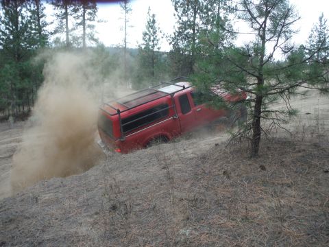 2005 Jeep Wrangler - A red pickup truck navigates a dusty, off-road trail, surrounded by trees and dry brush.