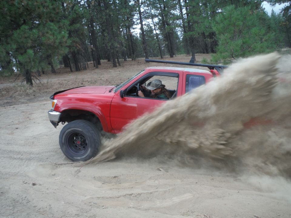 Red pickup truck driving through sand, kicking up a large dust cloud