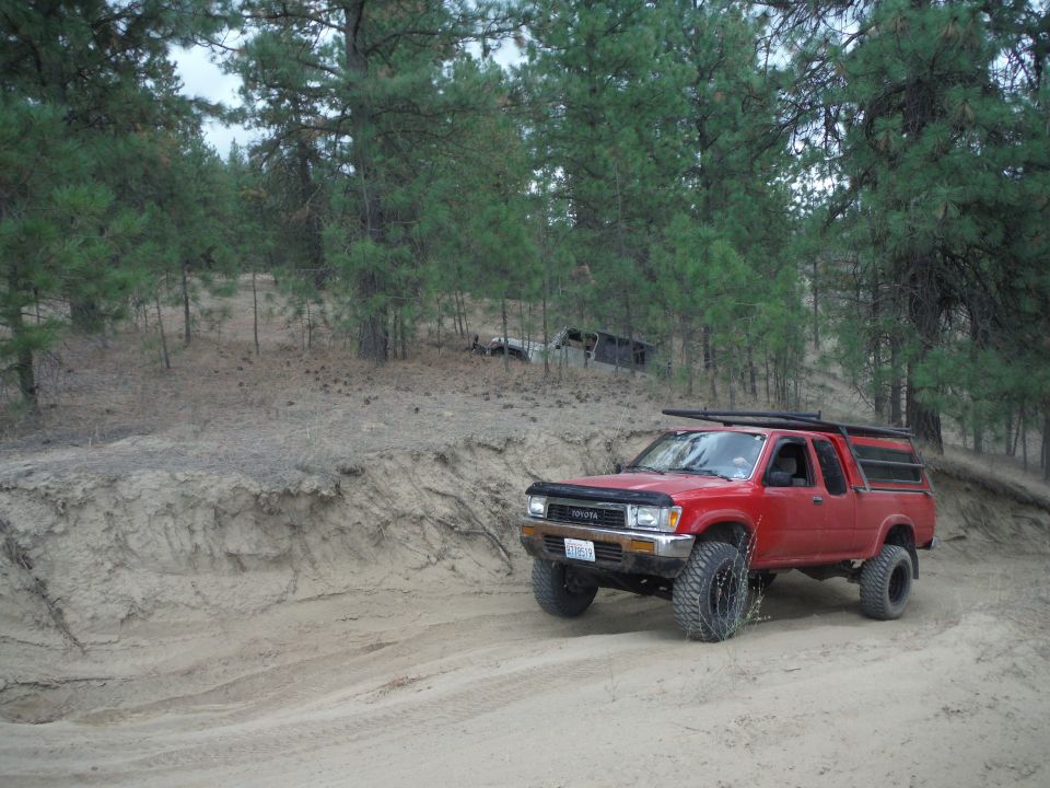 Red Toyota pickup truck with a camper shell driving on a dirt road, front 3/4 view. Modified wheels and tires