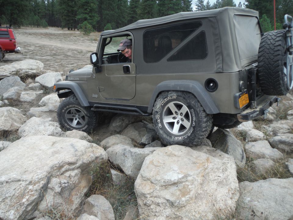 Olive green Jeep Wrangler TJ on rocks, driver visible, soft top, spare tire on rear