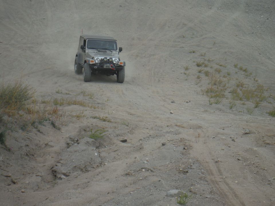 Silver Jeep Wrangler driving up a dirt hill, kicking up dust. It has a winch on the front