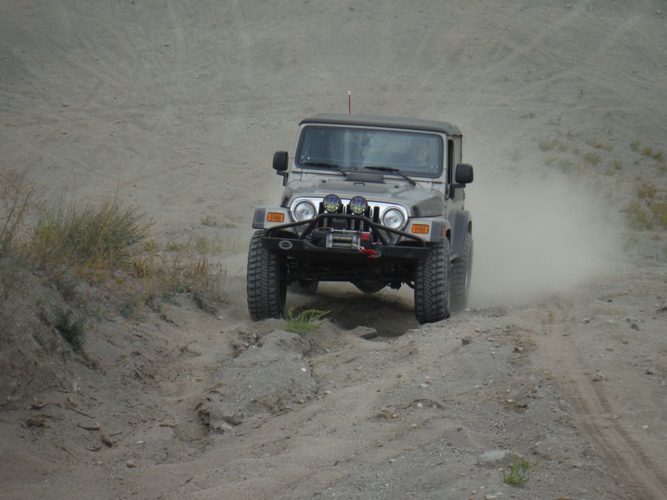 Silver Jeep Wrangler driving on a dirt road, kicking up dust. It has a winch and auxiliary lights on the front bumper