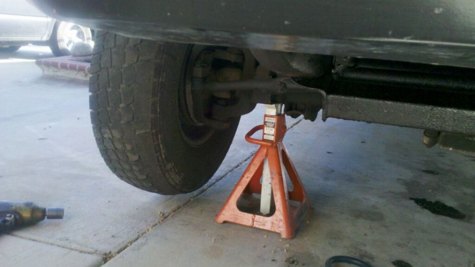 Truck chassis with front wheel elevated on an orange jack stand, showing suspension components and a black frame rail