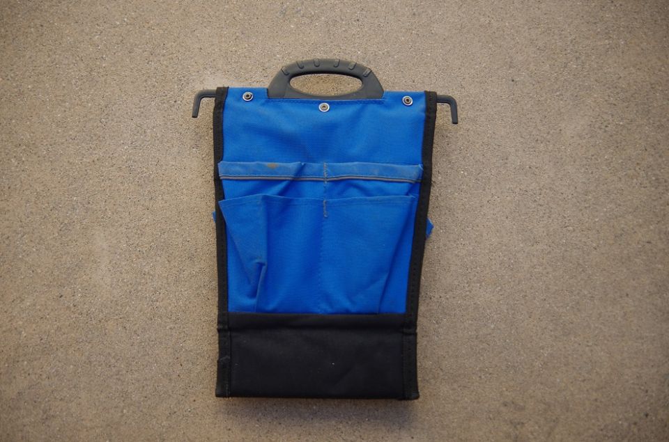 Blue fabric tool roll organizer with black trim and handle, hanging on a textured wall