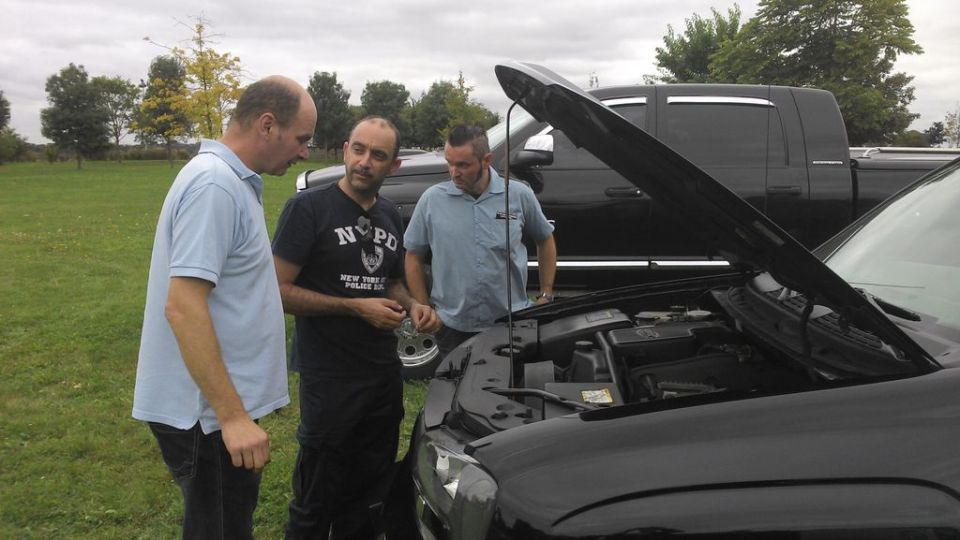 Three men examine the engine bay of a black car with its hood open; a black pickup truck is parked behind it