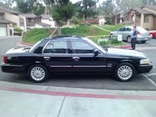 2011 Mercury Grand Marquis - A black sedan is parked on a street with a person standing near another vehicle in the background.