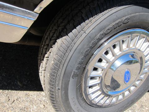 1991 Ford LTD Crown Victoria - Close-up of a vehicle's front wheel showing a Firestone tire and chrome alloy hubcap with a blue Ford emblem at its center.