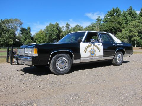 1991 Ford LTD Crown Victoria - black-and-white Ford Highway Patrol sedan with a front push bar parked on gravel before trees under a blue sky.