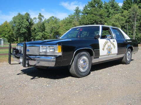 1991 Ford LTD Crown Victoria - Front three-quarter view of a black-and-white Highway Patrol sedan with quad headlights and a front push bar, parked on gravel.