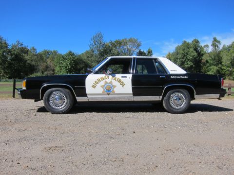 1991 Ford LTD Crown Victoria - Side profile of a black-and-white four-door Highway Patrol sedan with the gold star emblem, parked on gravel under a clear blue sky.