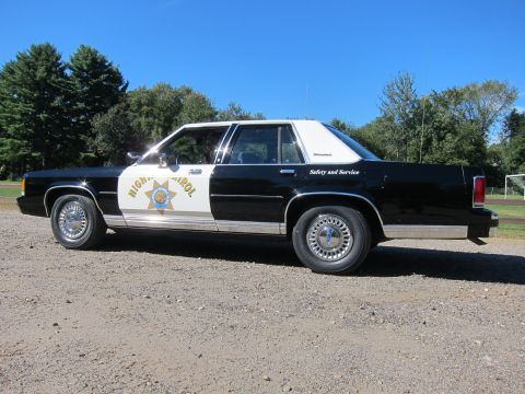 1991 Ford LTD Crown Victoria - Side profile of a black-and-white Highway Patrol sedan lettered Safety and Service, parked on gravel with trees in the background.