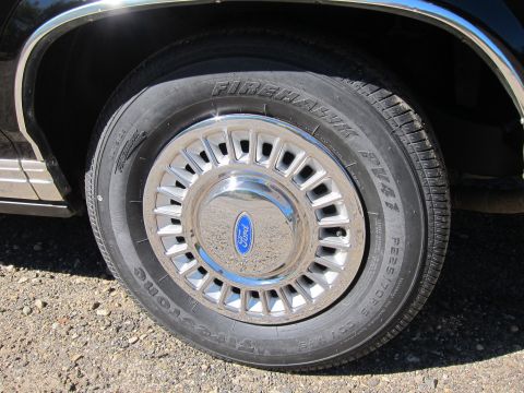 1991 Ford LTD Crown Victoria - Close-up of a black car's front wheel showing a chrome hubcap with the blue Ford oval logo mounted on a Firestone Firehawk tire.