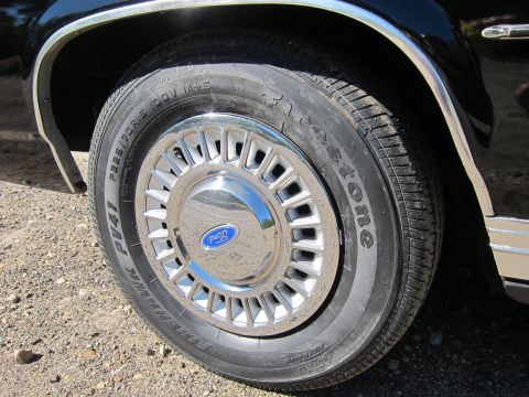1991 Ford LTD Crown Victoria - Close-up of a black car's wheel with a chrome Ford-logo hubcap mounted on a Firestone tire over gravel.