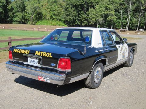1991 Ford LTD Crown Victoria - Rear three-quarter view of a black-and-white police sedan with roof emergency lights and yellow 'Highway Patrol' lettering on the trunk.