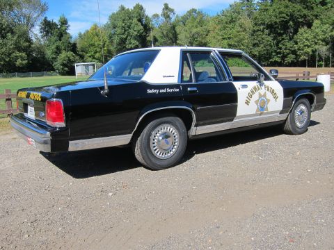1991 Ford LTD Crown Victoria - Rear three-quarter passenger-side view of a black-and-white police sedan in CHP livery with the gold star door emblem, parked on gravel.