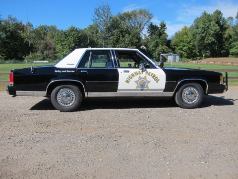 1991 Ford LTD Crown Victoria - Full passenger-side profile of a black-and-white police sedan in CHP livery, with whip antenna and gold 'Highway Patrol' star emblem on the…