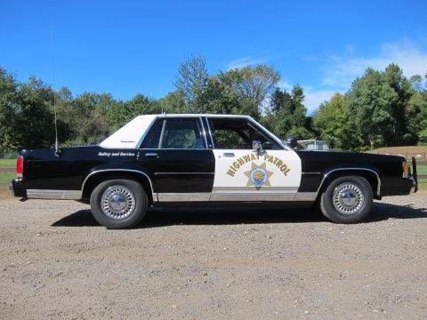 1991 Ford LTD Crown Victoria - Passenger-side profile of a black-and-white police sedan in California Highway Patrol livery, fitted with a front push bar and gold star do…