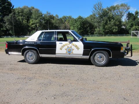 1991 Ford LTD Crown Victoria - Passenger-side profile of a black-and-white police sedan in CHP livery, with a whip antenna and front push bar, parked on gravel by a field.