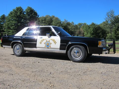 1991 Ford LTD Crown Victoria - Front three-quarter view of a black-and-white California Highway Patrol full-size sedan with push bar, sun glaring off the windshield.