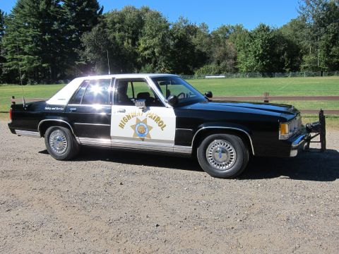 1991 Ford LTD Crown Victoria - Driver-side three-quarter view of a black-and-white CHP-liveried full-size Ford sedan parked on gravel near a field.