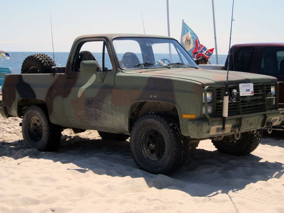 Camouflaged pickup truck with spare tire in bed, parked on a beach with fishing rods in the background
