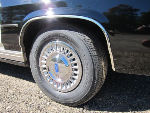 1991 Ford LTD Crown Victoria - Close-up of a black sedan's front wheel with a chrome turbine-style Ford hubcap and a Firestone whitewall tire.