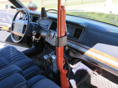 1991 Ford LTD Crown Victoria - Interior of a Ford police car showing a vertically mounted shotgun, an in-dash two-way radio head, and a blue cloth front bench seat.