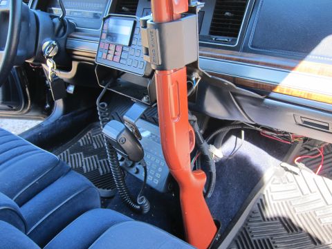 1991 Ford LTD Crown Victoria - Close-up of a shotgun secured in a vertical floor mount next to a two-way radio control head and handheld microphone inside a police car.