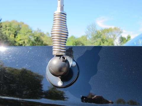 1991 Ford LTD Crown Victoria - chrome coil-spring antenna base mounted on the black bodywork of a car, set against a clear blue sky and trees.