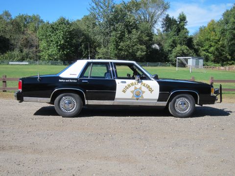 1991 Ford LTD Crown Victoria - Driver-side profile of a black-and-white Highway Patrol Ford sedan showing the door shield emblem and roof antennas, parked by a field.