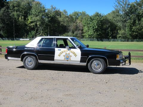 1991 Ford LTD Crown Victoria - Passenger-side view of a black-and-white Highway Patrol Ford sedan with a front push bar and the patrol shield on the front door.