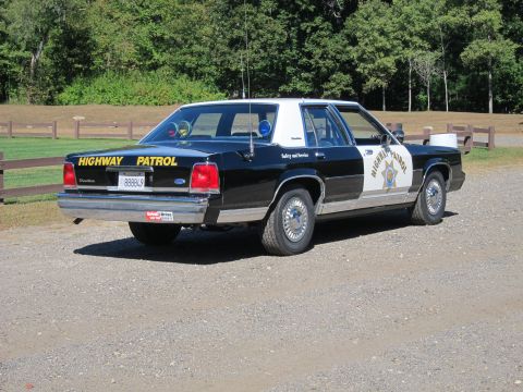 1991 Ford LTD Crown Victoria - Rear three-quarter view of a black-and-white Highway Patrol Ford sedan with yellow trunk lettering, the door shield, and a Ford trunk badge.