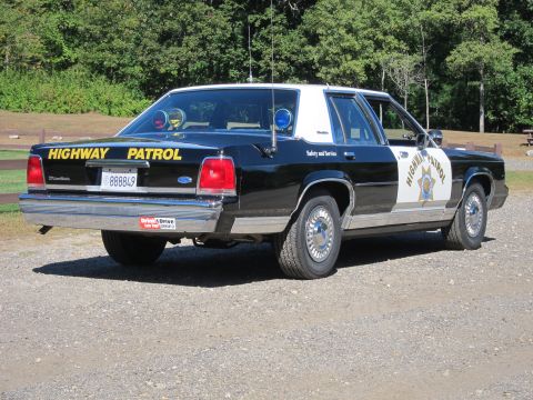 1991 Ford LTD Crown Victoria - Rear three-quarter view of a black-and-white Highway Patrol Ford sedan parked on gravel, showing the door shield and yellow trunk lettering.
