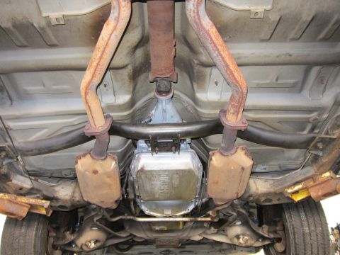 1991 Ford LTD Crown Victoria - Undercarriage view of a rear floor pan with rust-streaked dual exhaust pipes flanking a Ford-stamped transmission pan.