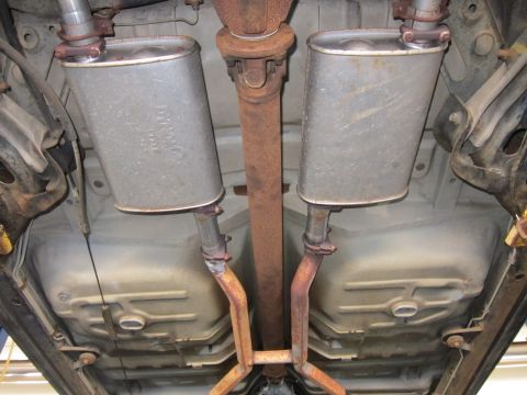 1991 Ford LTD Crown Victoria - Undercarriage view of twin oval mufflers, a rust-coated driveshaft and crossover exhaust pipe beneath a vehicle.