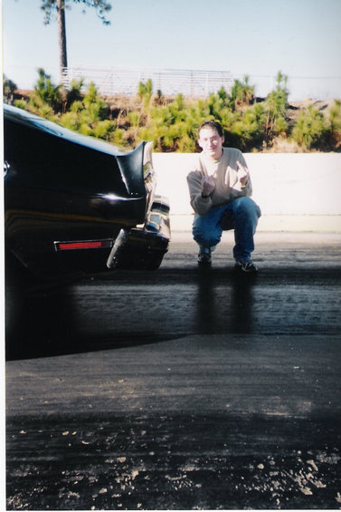 1977 Ford Mustang II - A person kneels beside a black car on a race track, giving a middle finger.