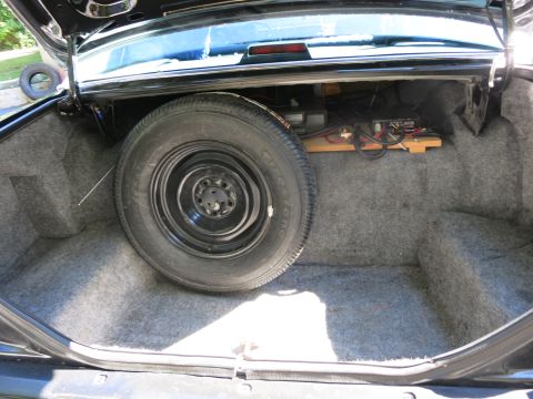 1991 Ford LTD Crown Victoria - Open carpeted car trunk showing a black steel spare wheel standing upright beside electronics on a wooden shelf.