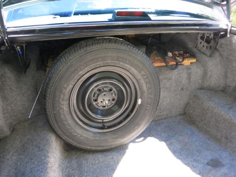 1991 Ford LTD Crown Victoria - Black steel spare wheel with a Firestone P225/70R15 tire standing in a carpeted car trunk beside wiring.