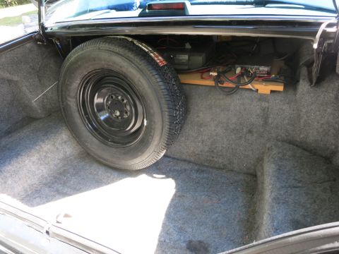 1991 Ford LTD Crown Victoria - Black steel spare wheel and tire standing in a carpeted car trunk beside electronics on a wooden shelf.