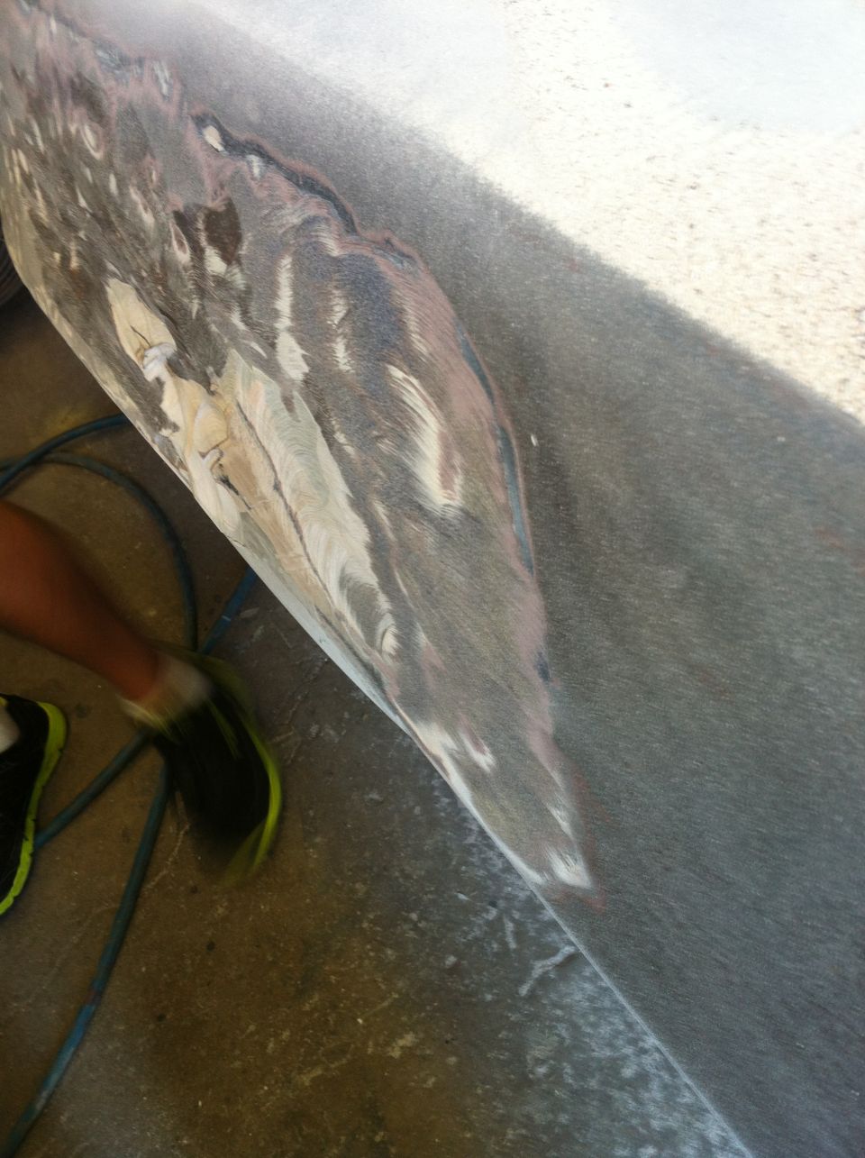 close-up of a car body panel with primer and sanding marks, a person's leg and shoe visible on the left